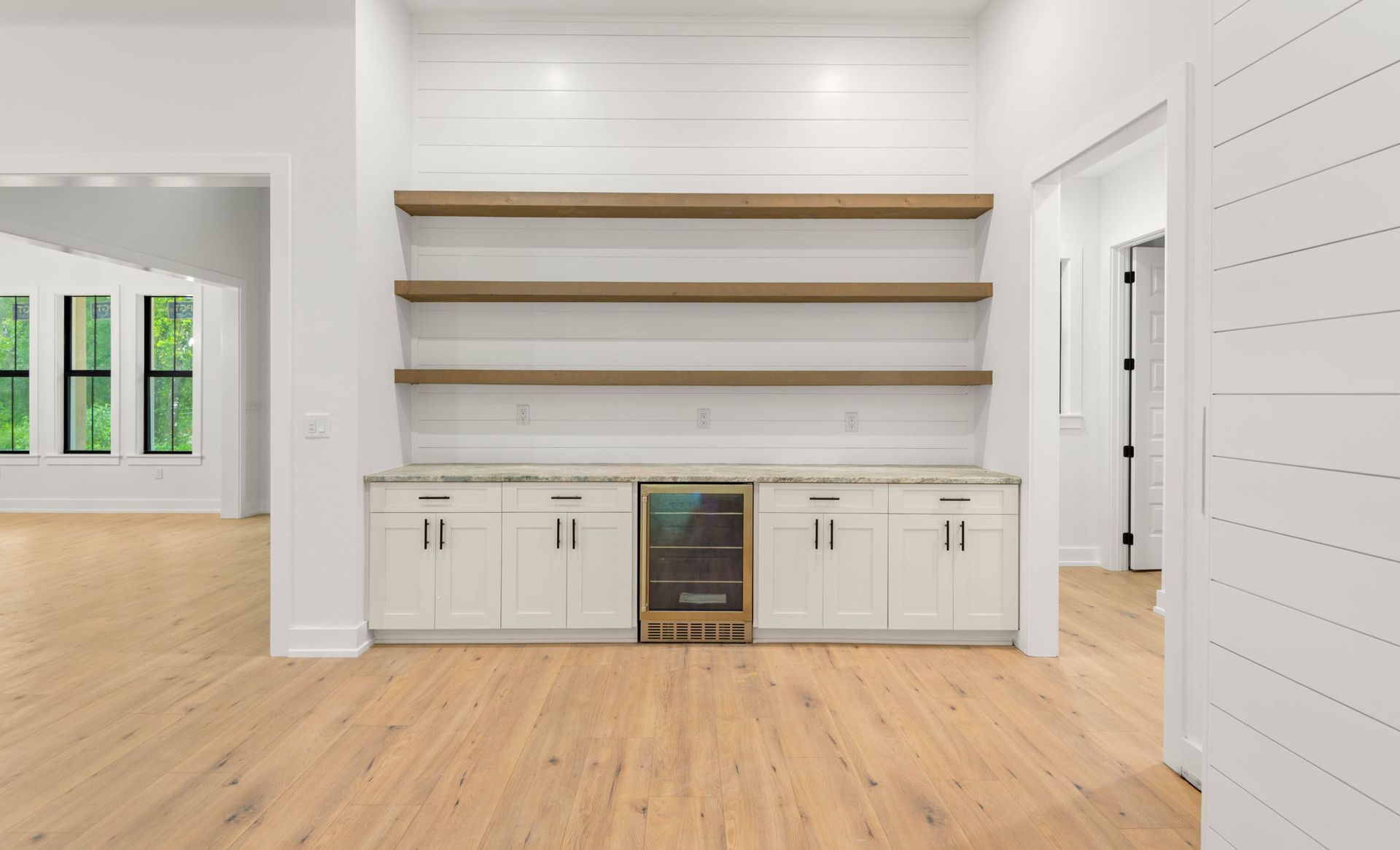 Built-in home bar with white cabinets, wood shelves, and a mini-fridge against a white shiplap wall.
