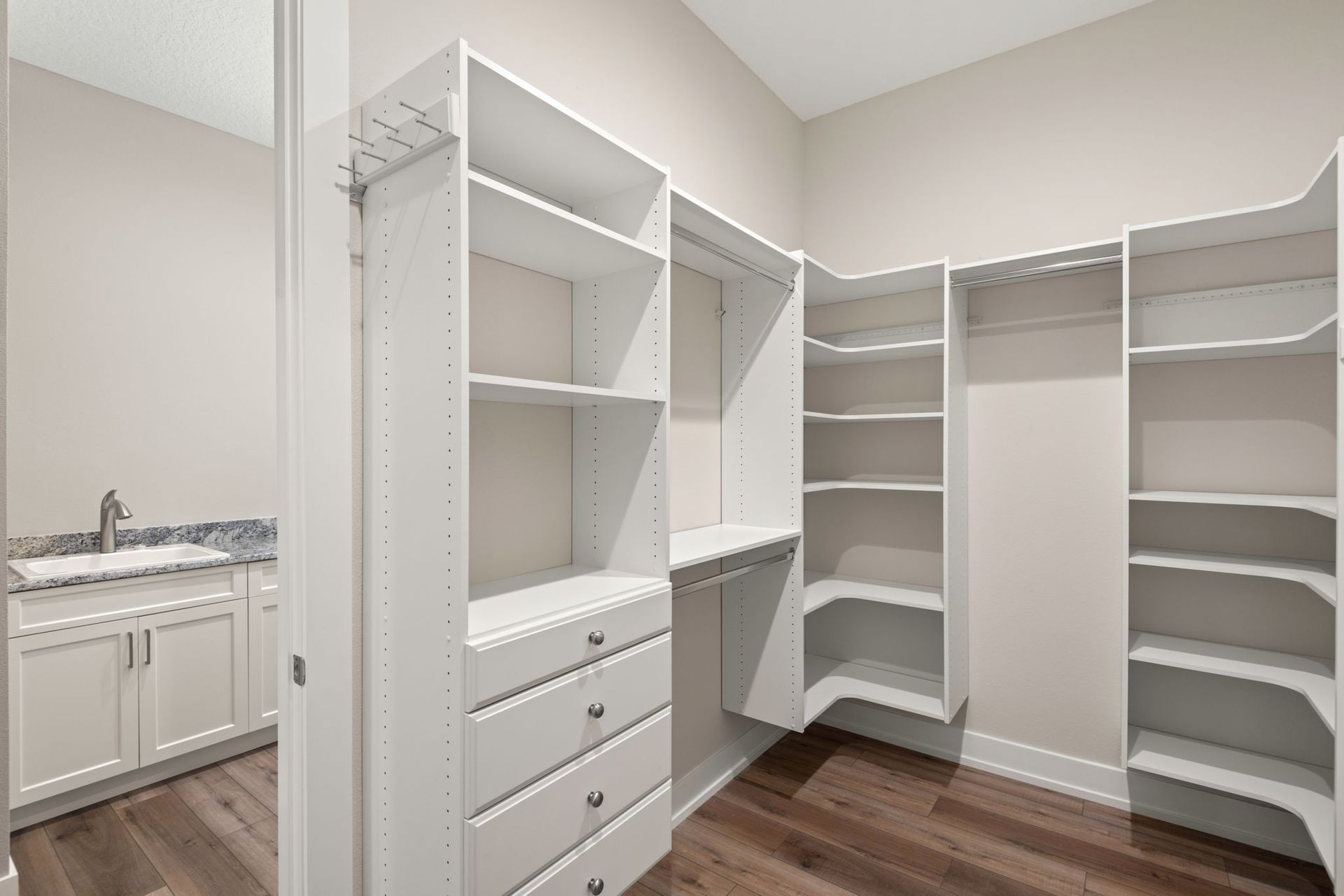 Walk-in closet with white shelves, drawers, and a vanity with a sink. Wooden floor and light walls.