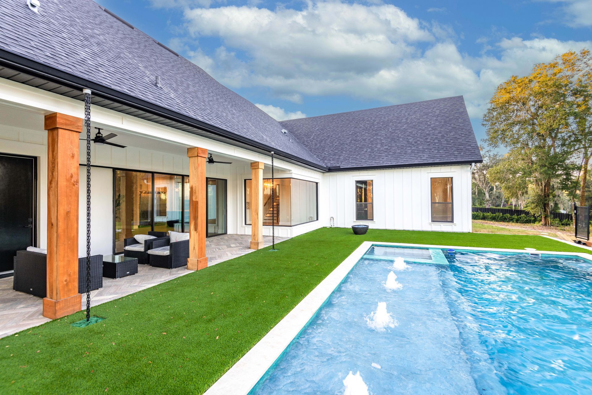 White house with pool. Artificial turf, blue water, wooden pillars, and a cloudy sky.