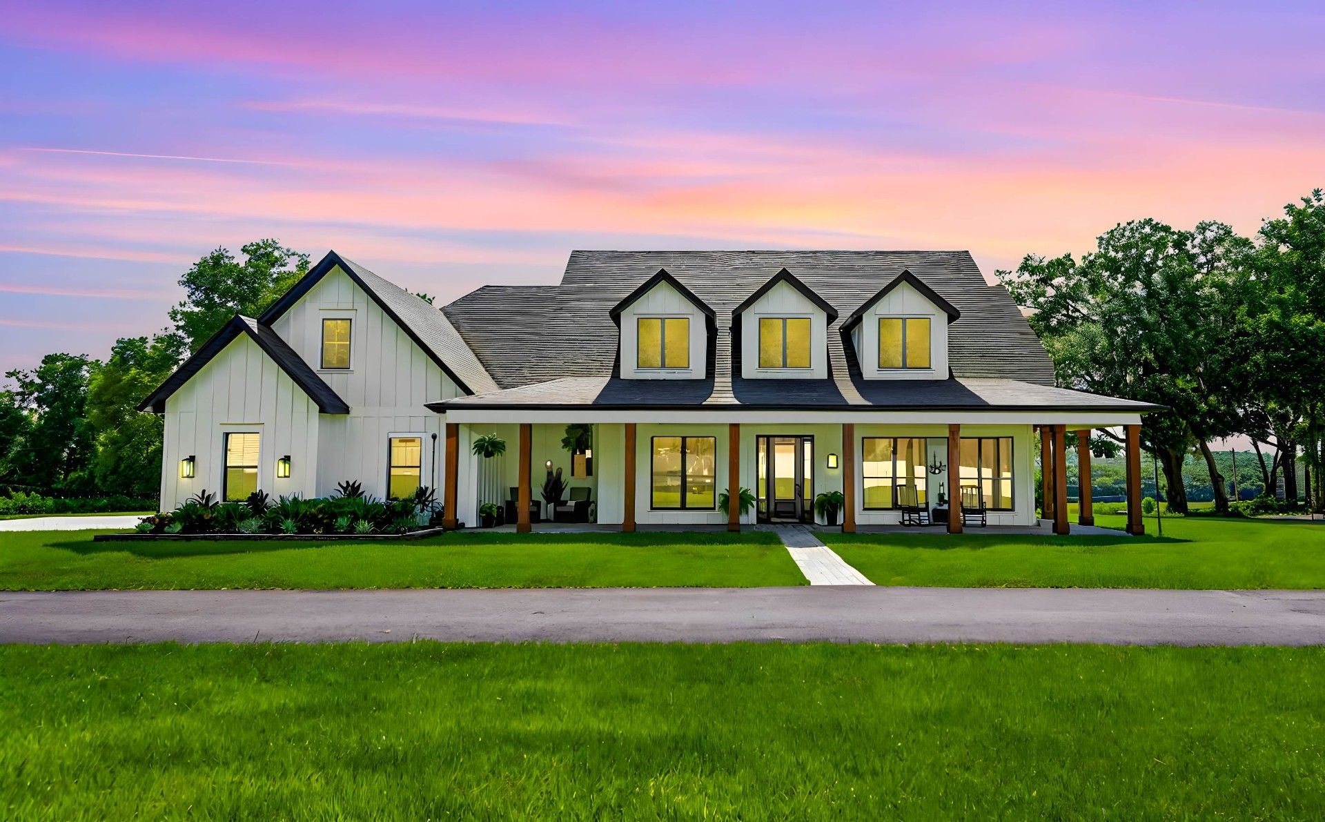 White farmhouse with black trim, porch, and dormers, set against a green lawn and vibrant sunset.