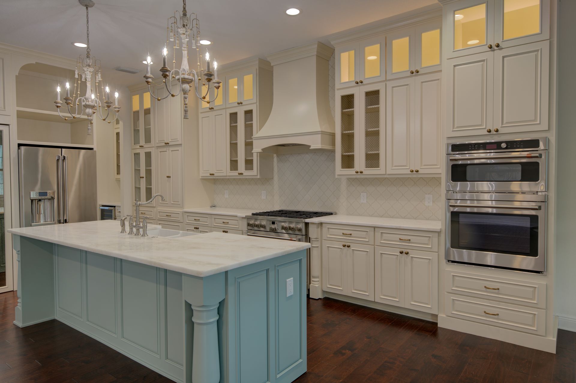 Elegant white kitchen with a blue island, chandeliers, stainless steel appliances, and dark wood floors.
