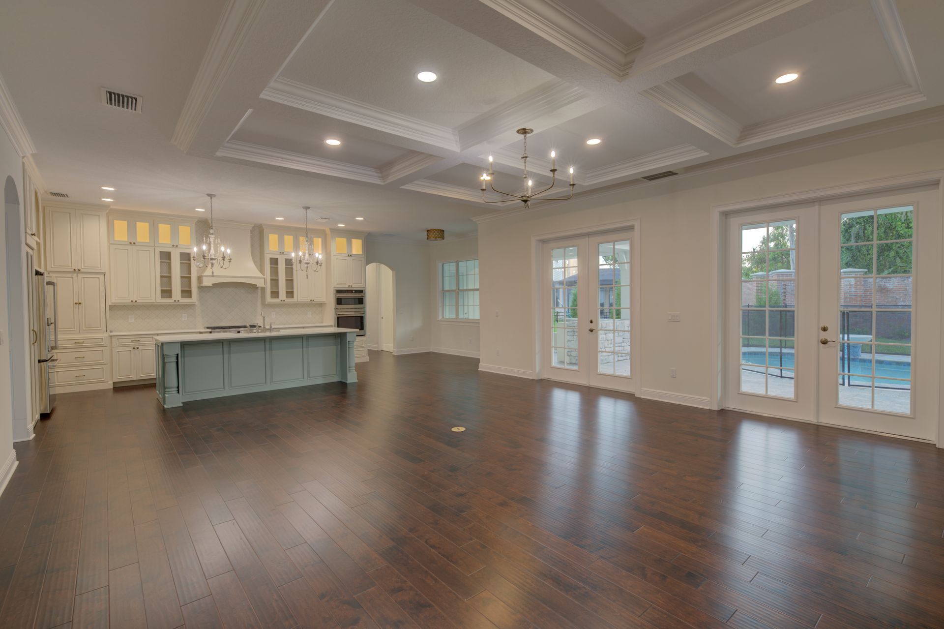 Spacious, empty room with a dark wood floor, kitchen, and pool view.