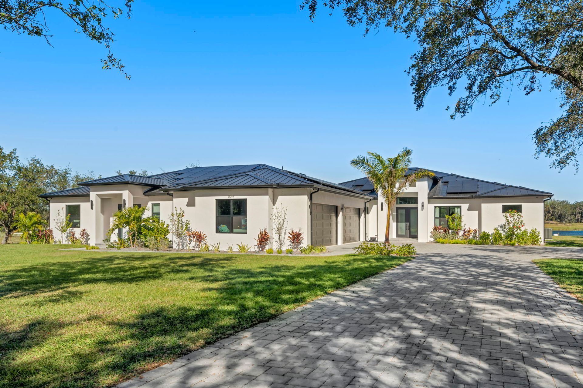 Modern, white stucco house with dark roof, long driveway, and green lawn under a blue sky.