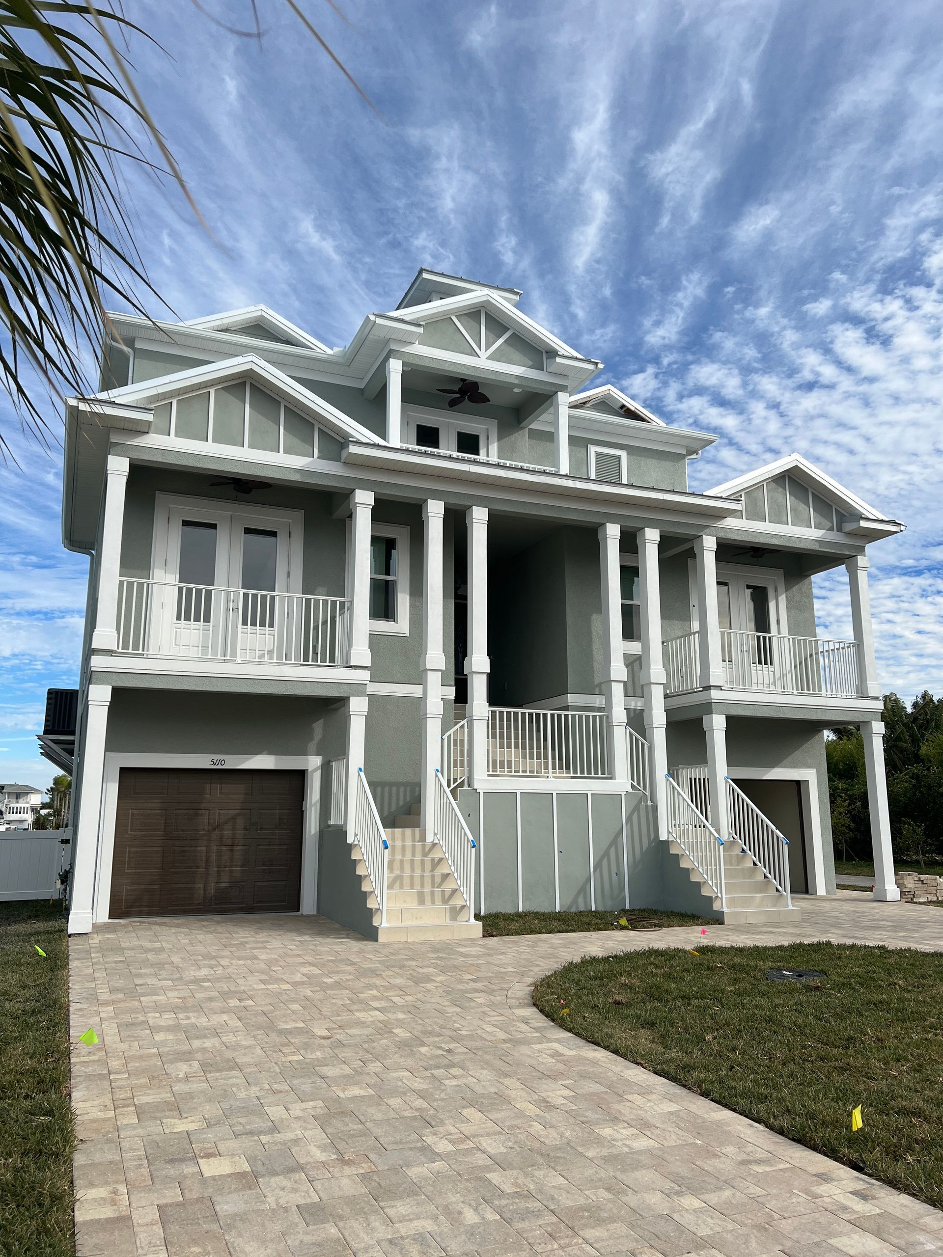 Three-story light blue house with white trim, balconies, and a brick driveway under a cloudy sky.