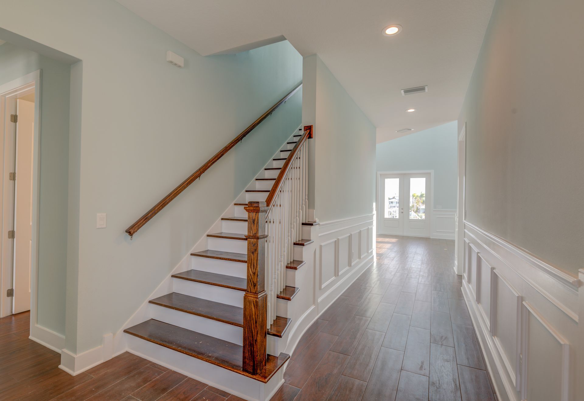 Interior hallway with staircase, pale blue walls, wood floors and trim, and white wainscoting.