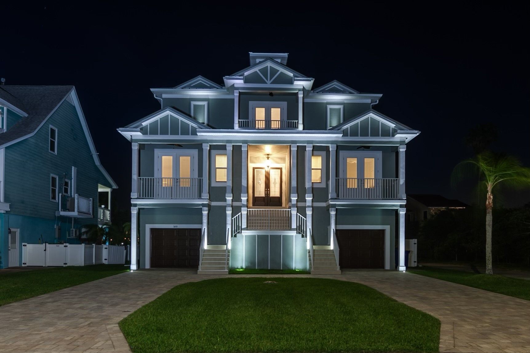 Multi-story teal house at night with bright exterior lights and a brick driveway.