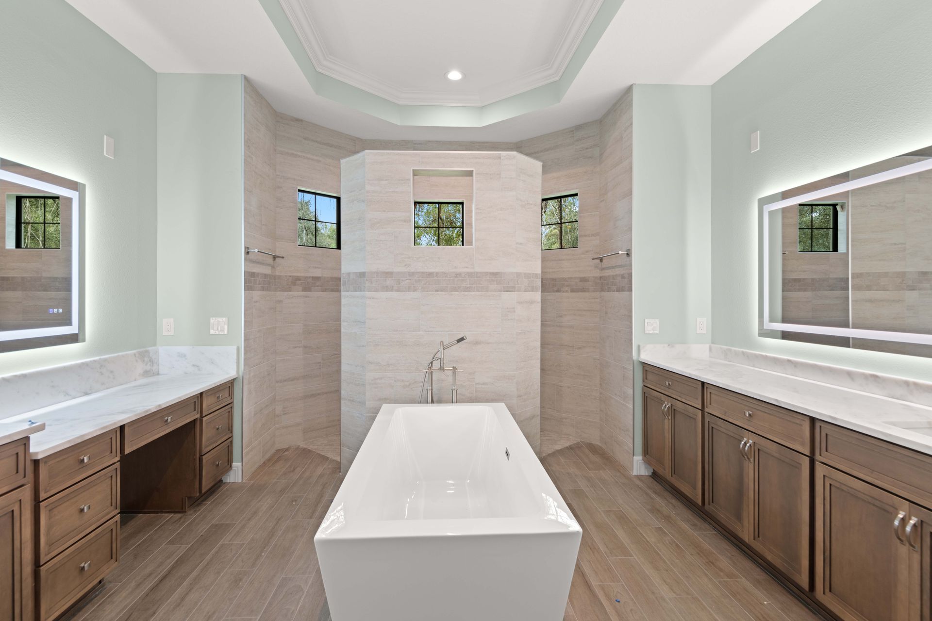 Spacious bathroom with light wood cabinets, a center tub, and light green walls. Two illuminated mirrors flank a shower.