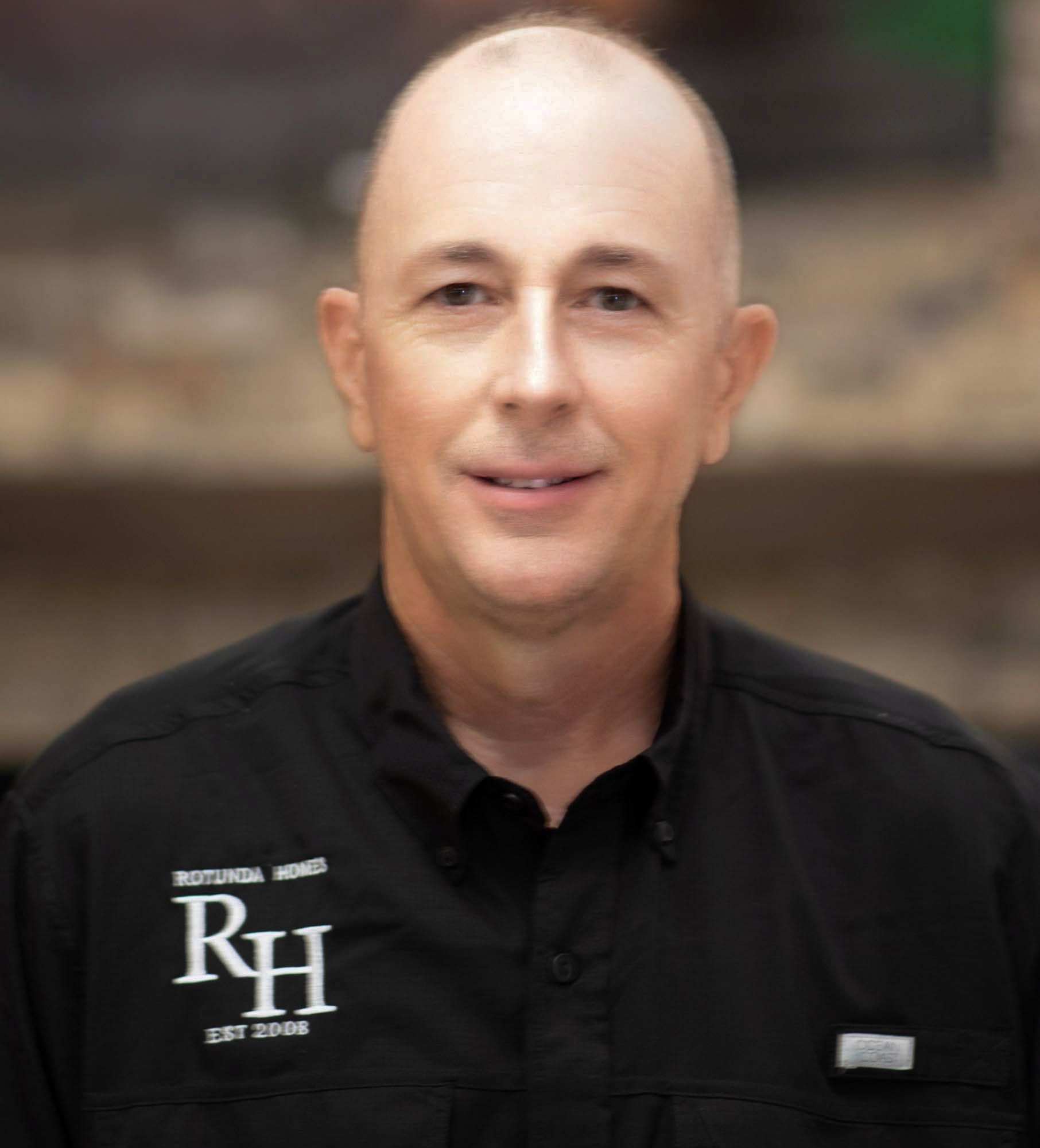 Bald man in black shirt with logo, smiling, indoors.