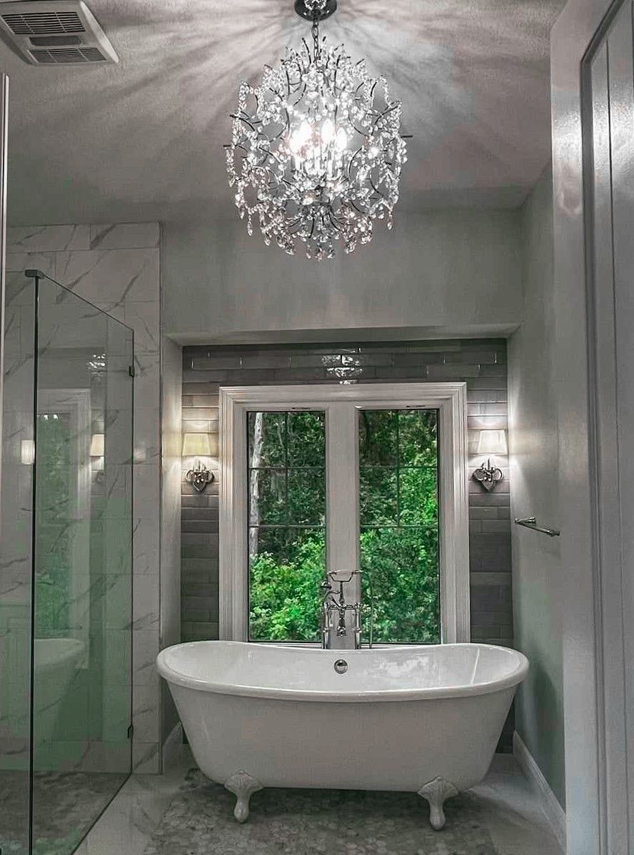 A clawfoot bathtub sits beneath a window with lush greenery, illuminated by a crystal chandelier in a tiled bathroom.