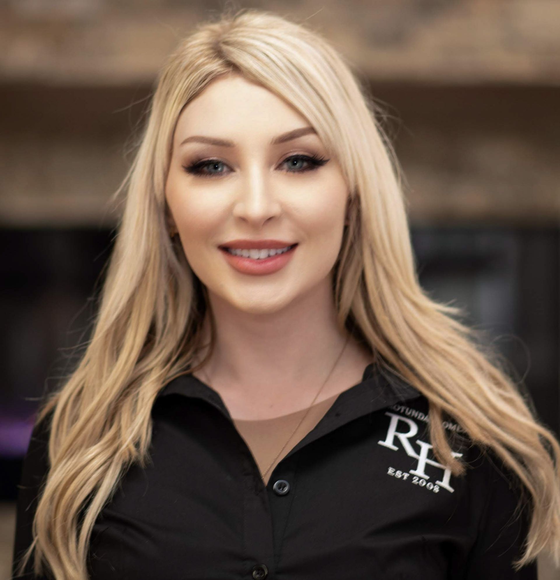 Blonde woman smiling, wearing a black shirt with a logo; indoors with a blurry background.
