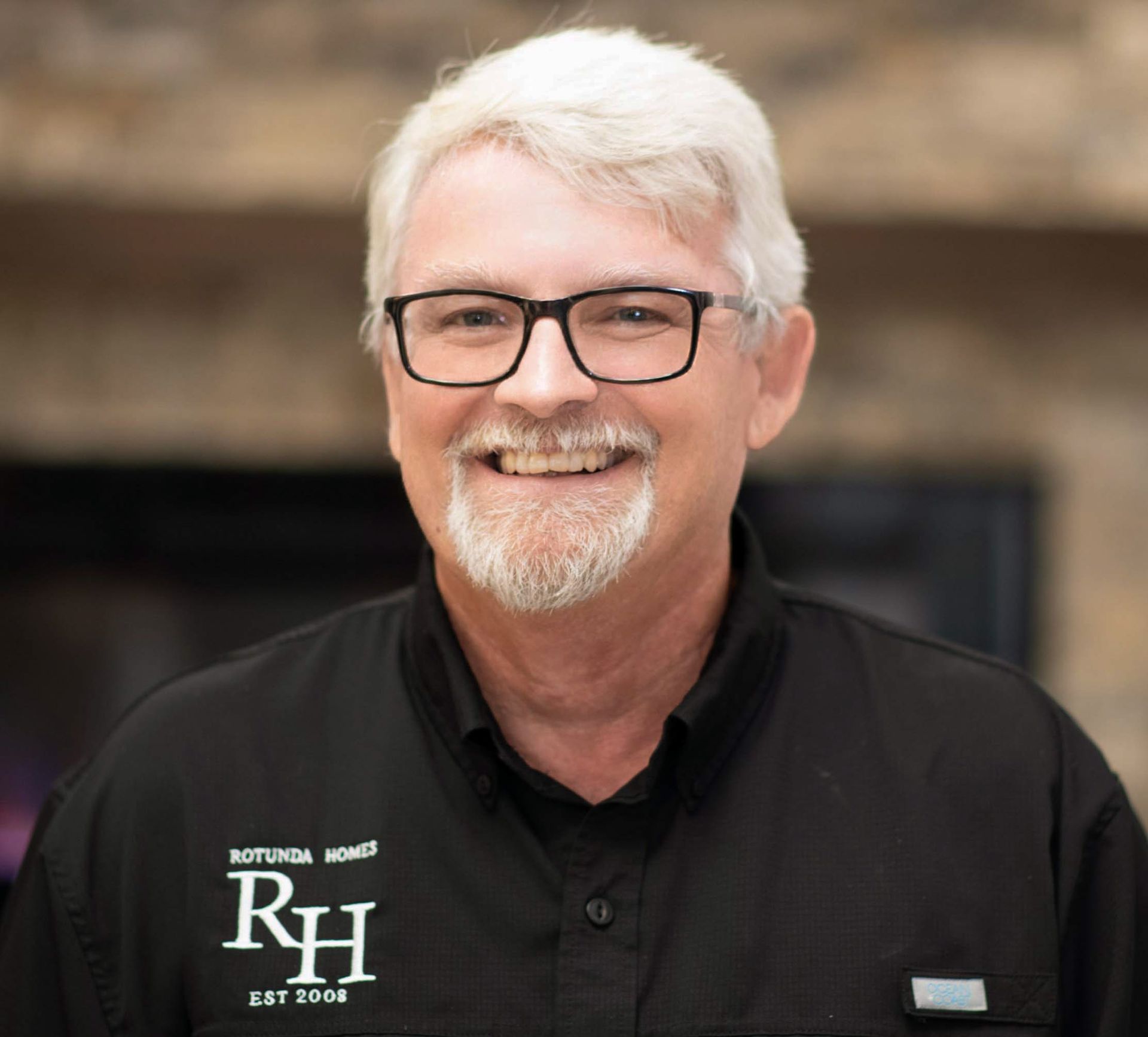Man with glasses smiling, wearing a black shirt with logo, in front of a fireplace.