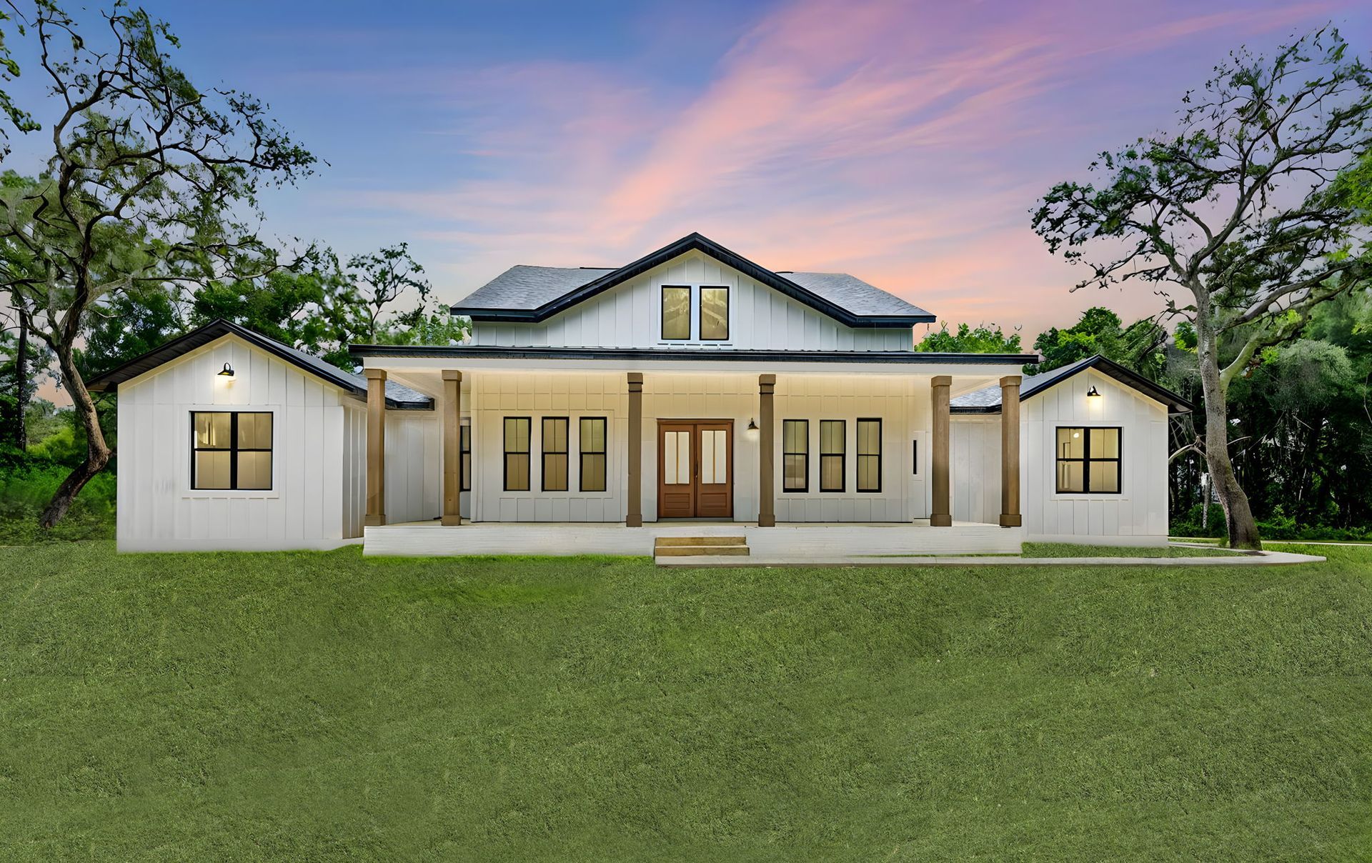 White farmhouse with a porch and two side buildings, set against a green lawn and a colorful sunset.