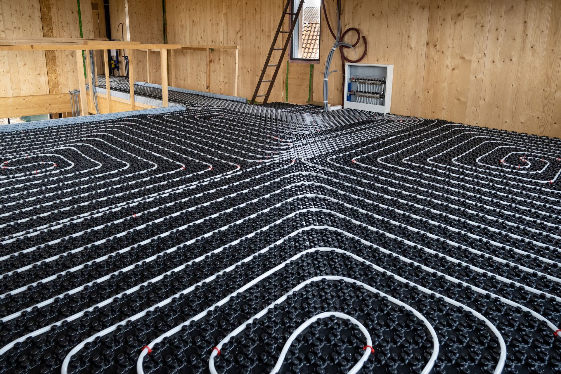 Tubi neri disposti a griglia sul pavimento, presumibilmente per il riscaldamento radiante, in una stanza con pareti in legno.