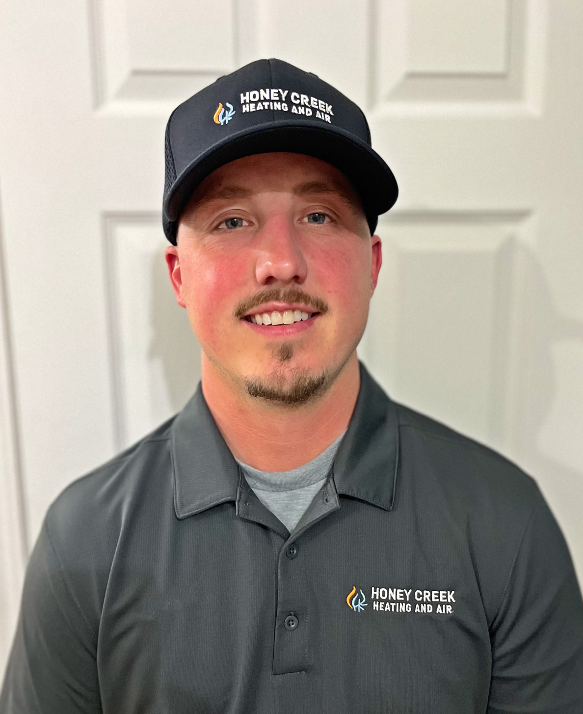 A smiling person wearing a dark grey company polo shirt and a matching baseball cap standing before a white door.
