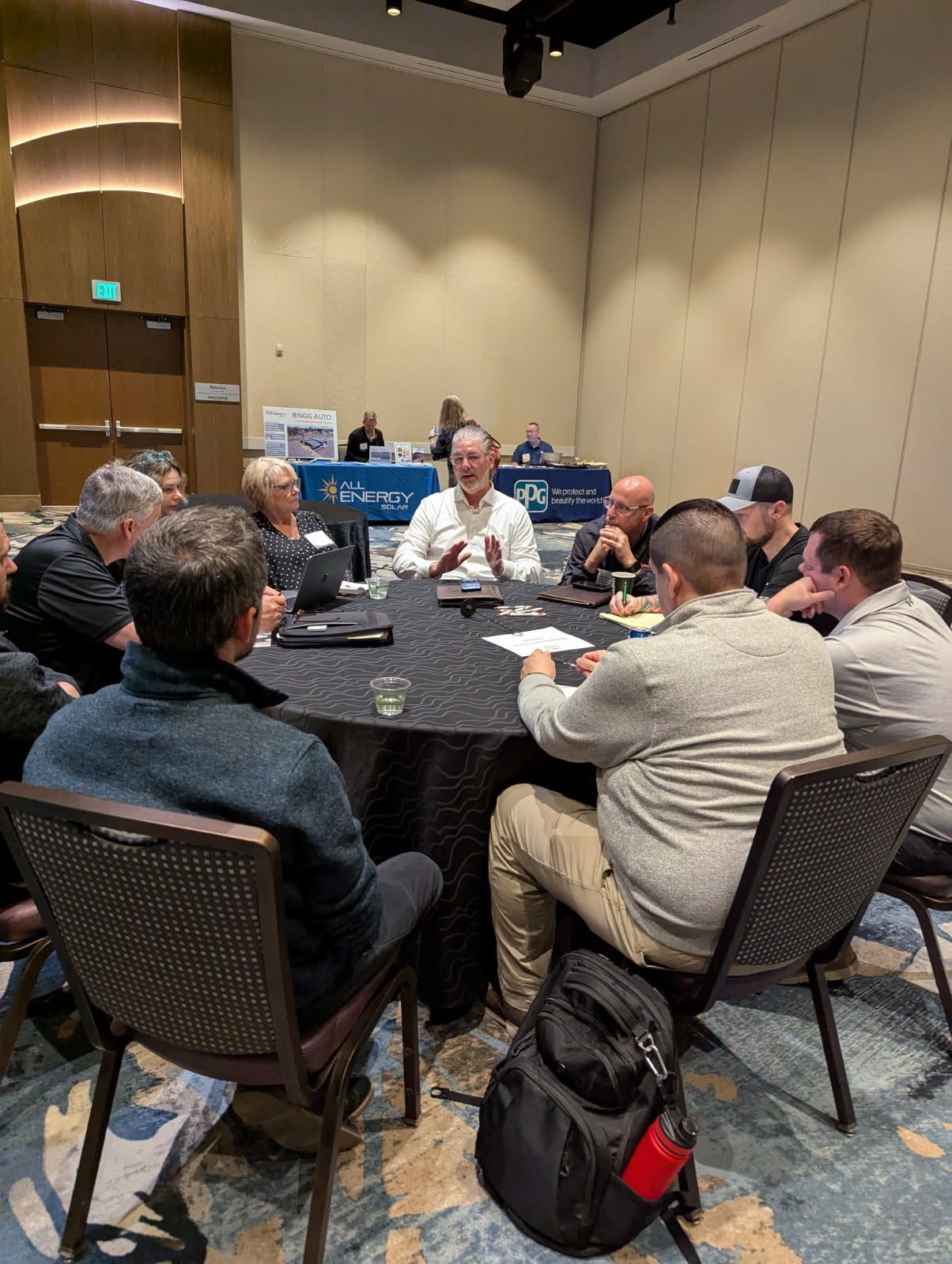 Group of people seated around a round table in a conference room. One person speaks.