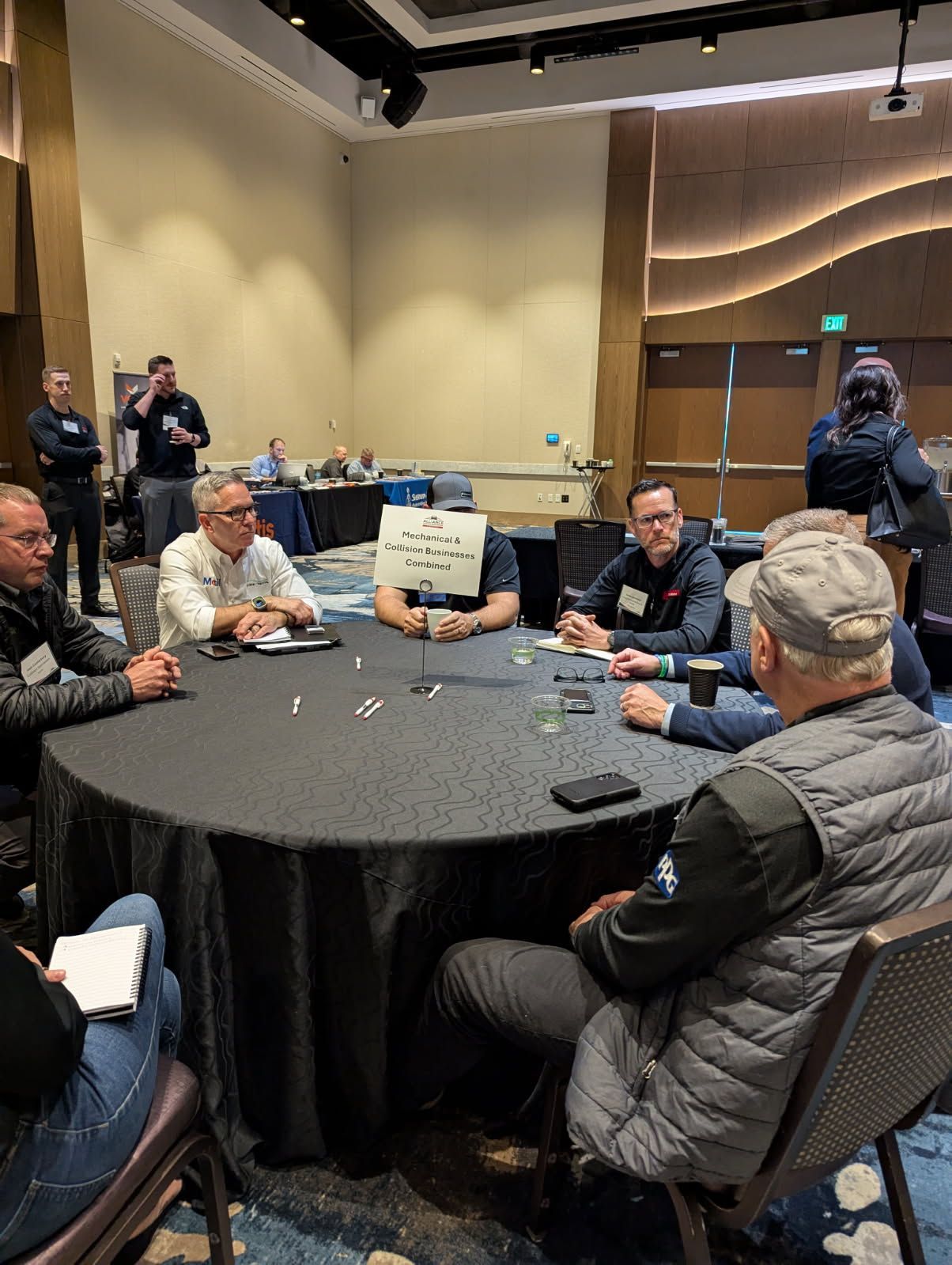 People sit around a round table in a conference room. Some are looking at the center of the table, and some are looking off.