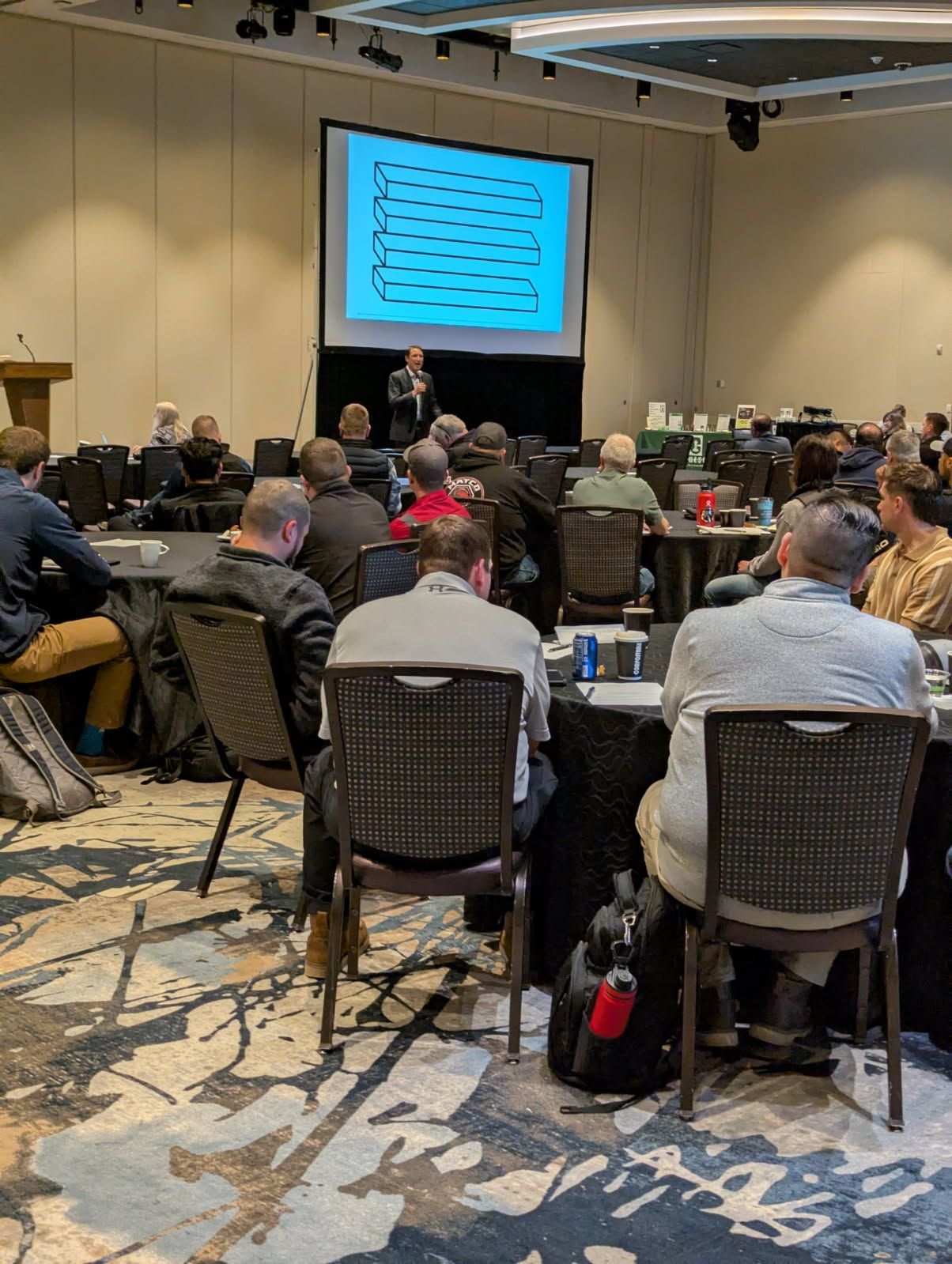A person gives a presentation in a conference room with an audience seated at tables. A screen displays text.