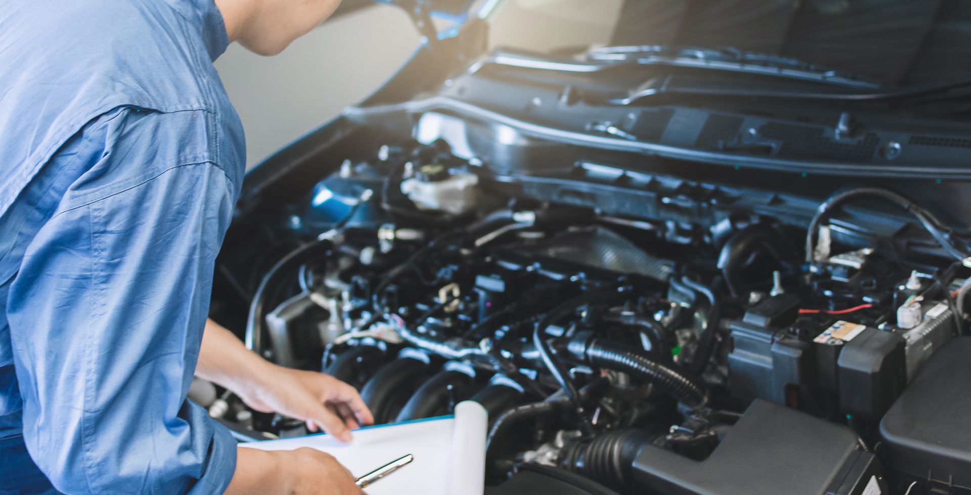 Mechanic inspecting car engine with a clipboard, blue uniform, indoor setting.