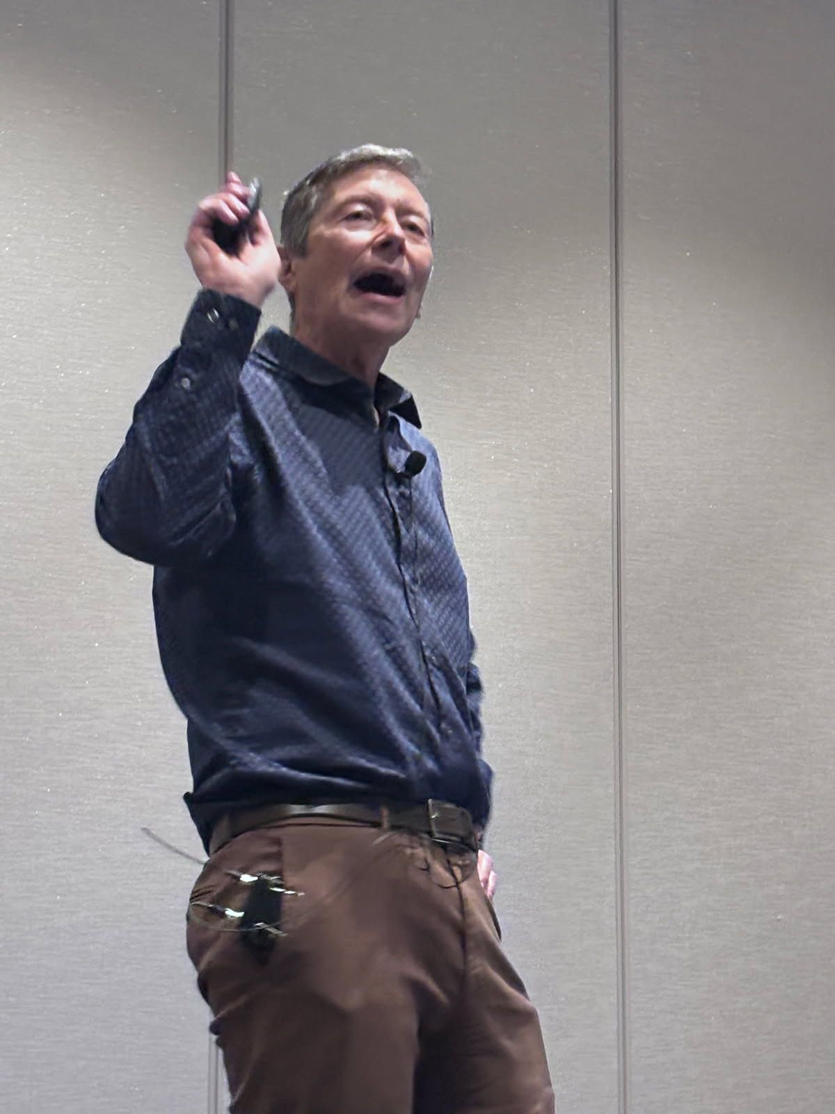 Man in blue shirt and brown pants gestures with a pen, speaking on a stage.