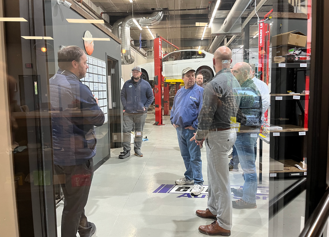 People in a workshop, looking at a whiteboard. Some wear work clothes, one looks at a car on a lift.