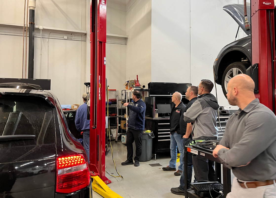 Group of men in a garage looking at a car on a lift; one points at something.