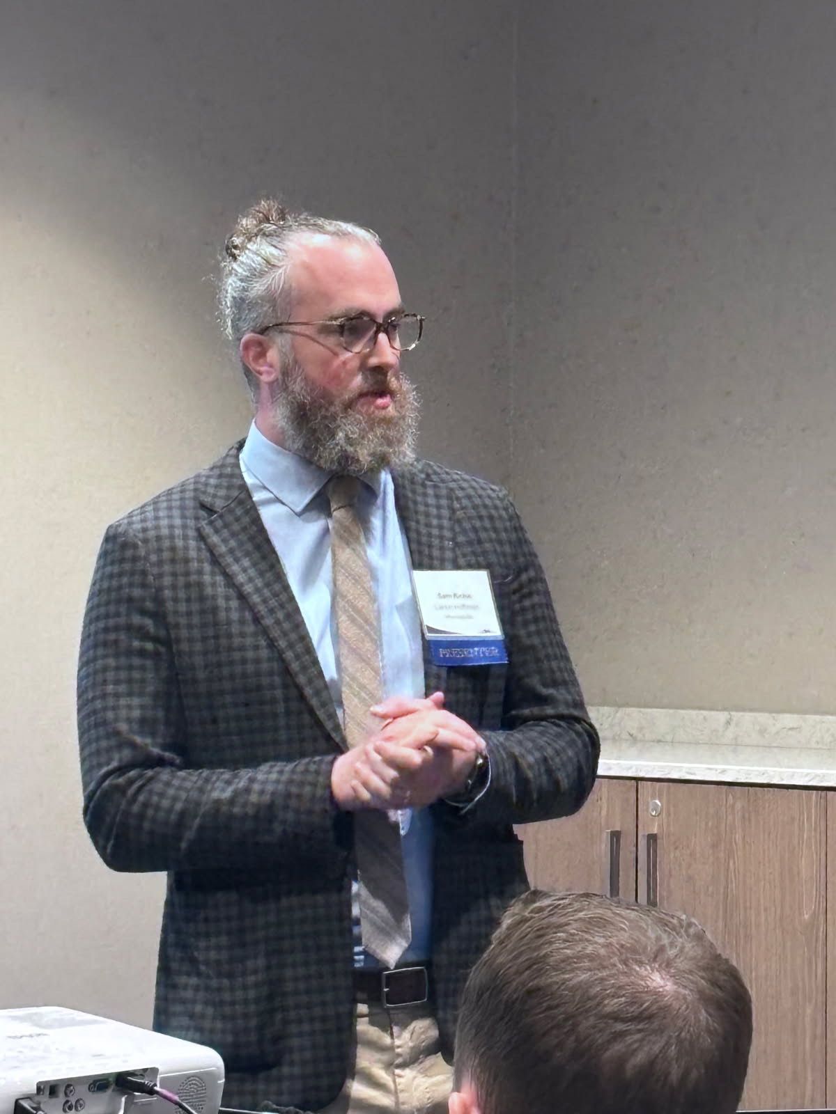 Man in blazer and tie speaking at a conference, hands clasped.