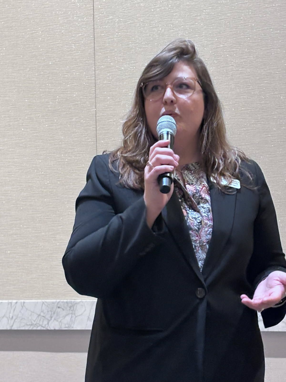 Woman in glasses and black blazer speaks into a microphone. Light-colored wall in the background.
