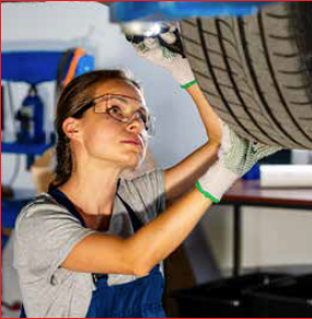 Woman in overalls, safety glasses, and gloves, examining a car tire in a garage.