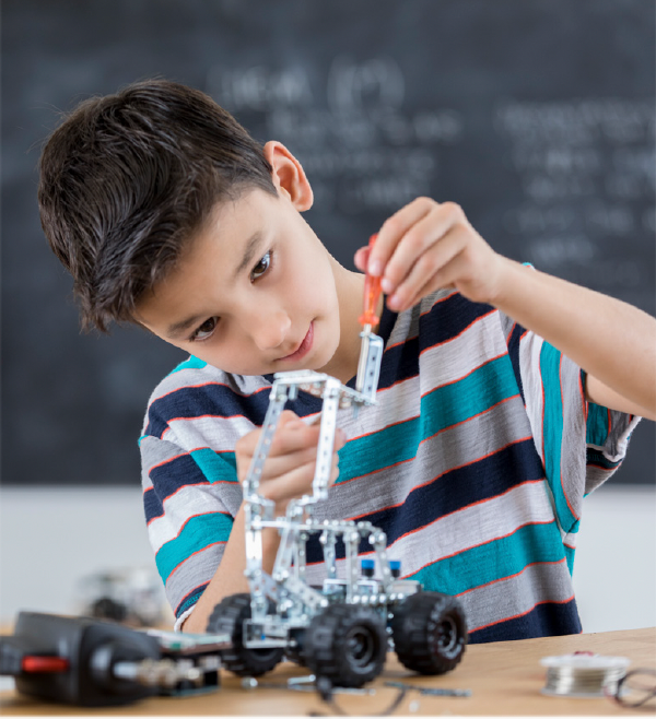Boy assembling a small robot with a screwdriver. Wooden table, chalkboard background.