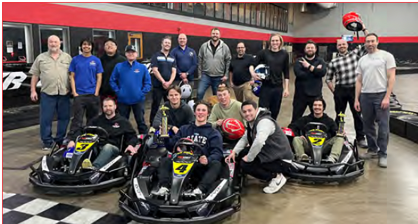 A group of people poses in an indoor go-kart facility after a race, with some seated in karts and others standing behind.