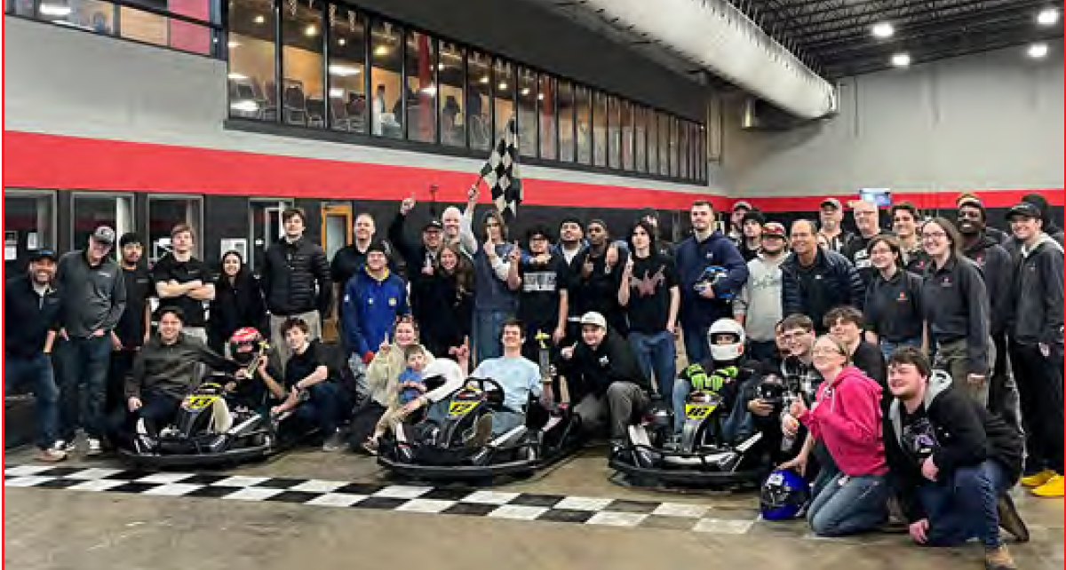 A large group of people posing together inside an indoor go-kart racing facility, some wearing helmets and seated in karts.