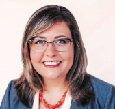 Woman wearing glasses smiles at the camera, wearing a blue blazer and orange necklace.
