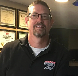 Man in glasses, black shirt, and gray undershirt smiles in a shop with framed certificates behind him.