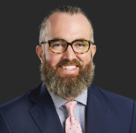 Man with a beard and glasses in a suit smiles at the camera against a dark background.