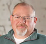 Headshot of a person with a goatee, wearing glasses and a dark green quarter-zip sweater against a neutral background.