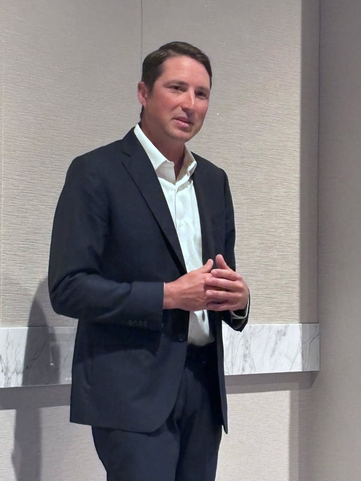 Man in a dark suit and open-collared shirt speaks at an event.