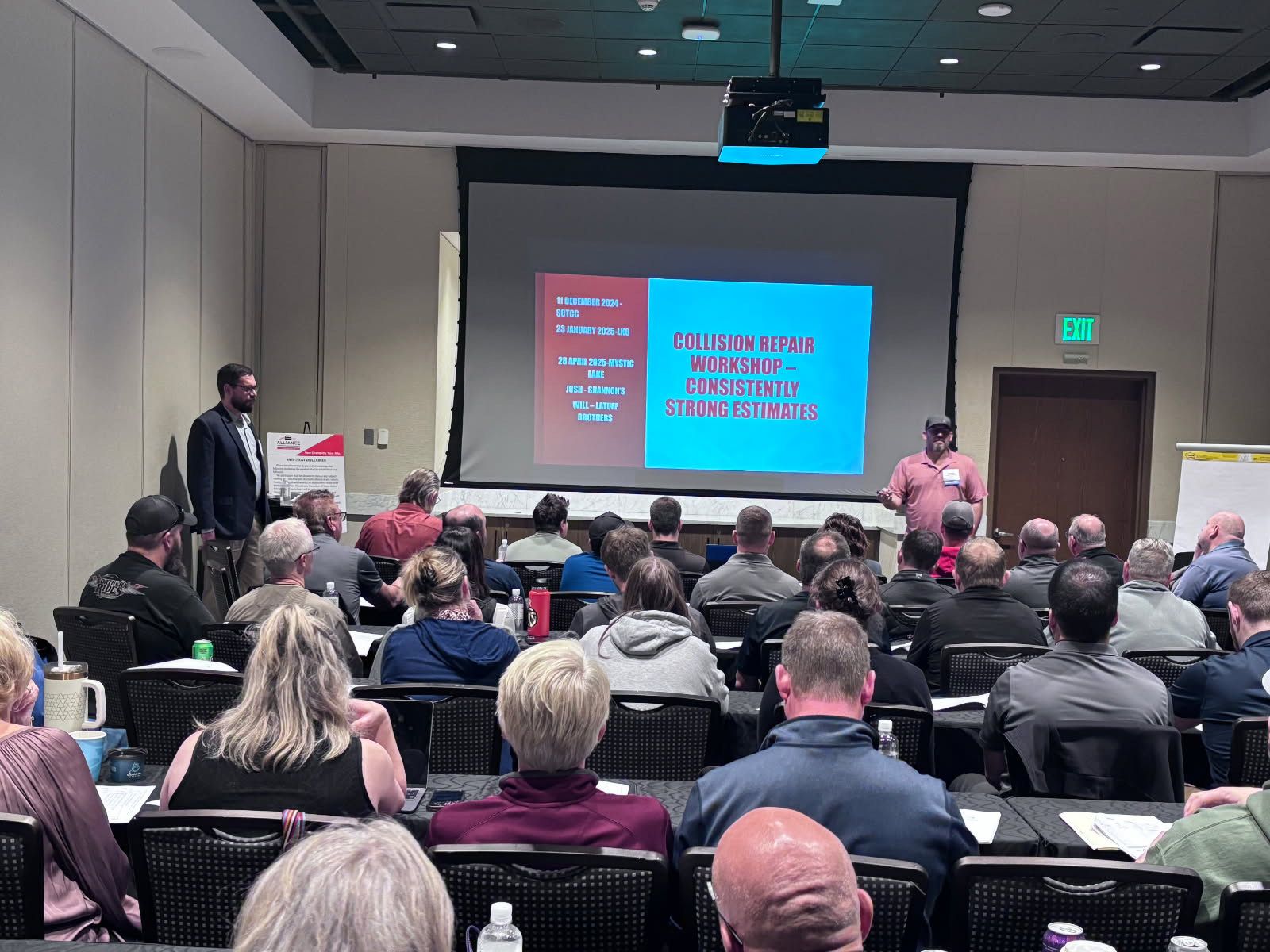 Presentation in a conference room. Two men stand before an audience, with a screen displaying text.