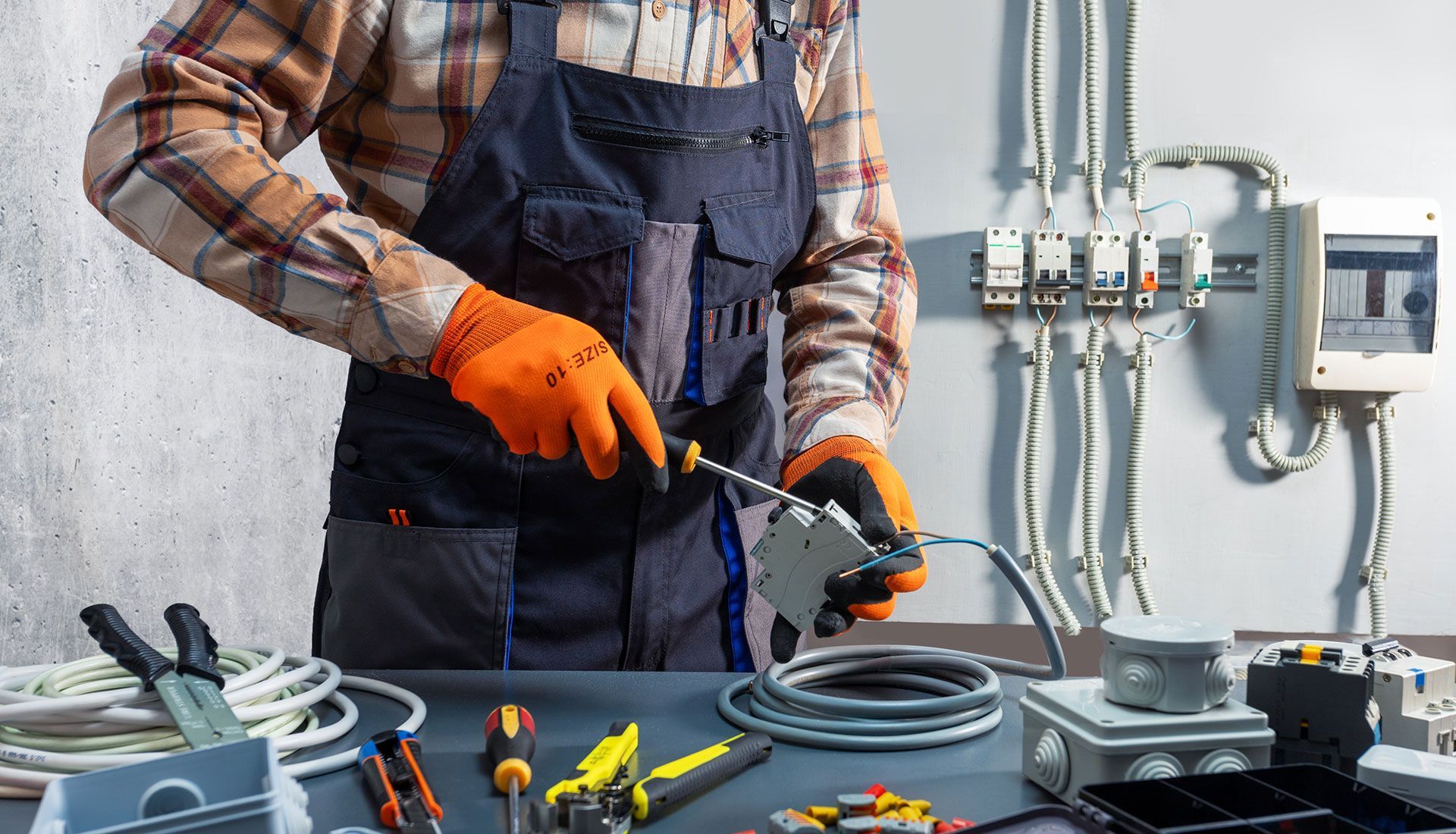 Electrician in workwear uses a screwdriver on a circuit breaker, surrounded by electrical tools and wiring on a workbench.