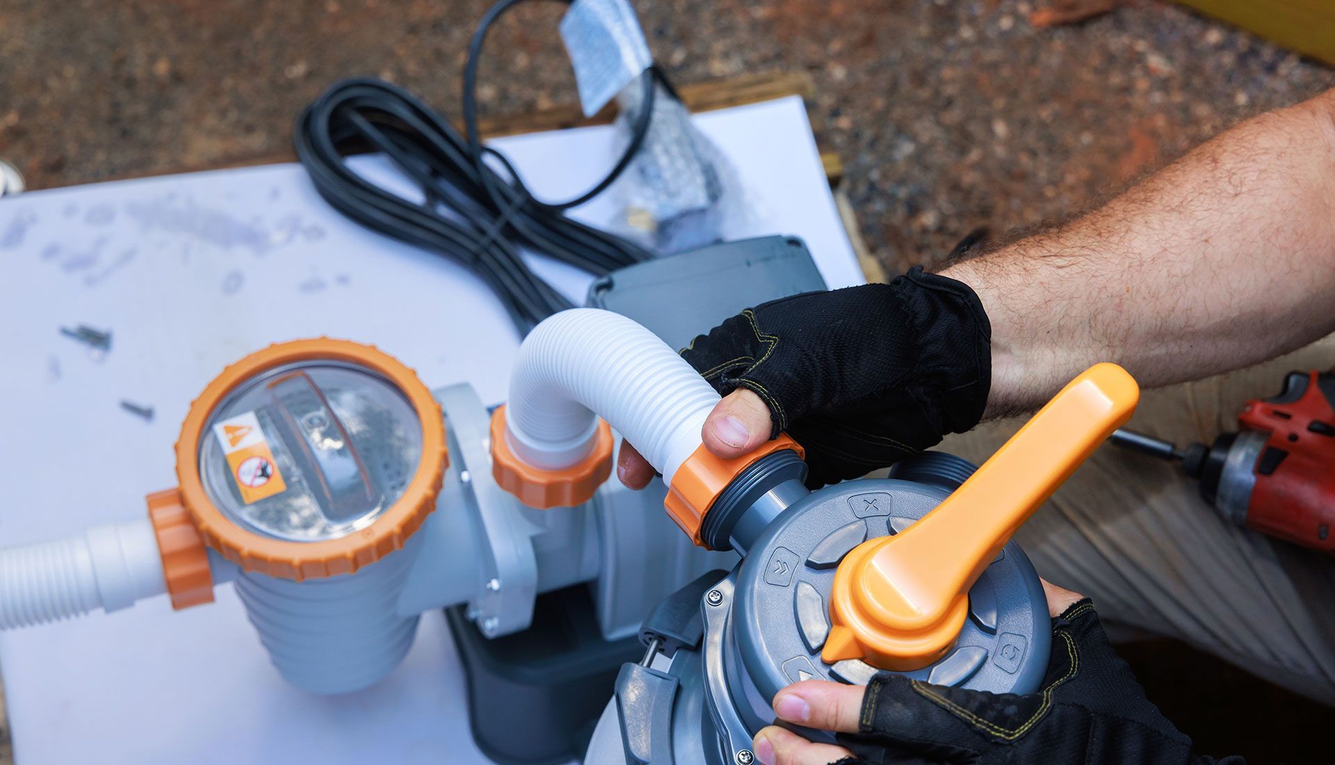 A person wearing gloves assembles a pool filter pump system, holding a grey valve with an orange handle.