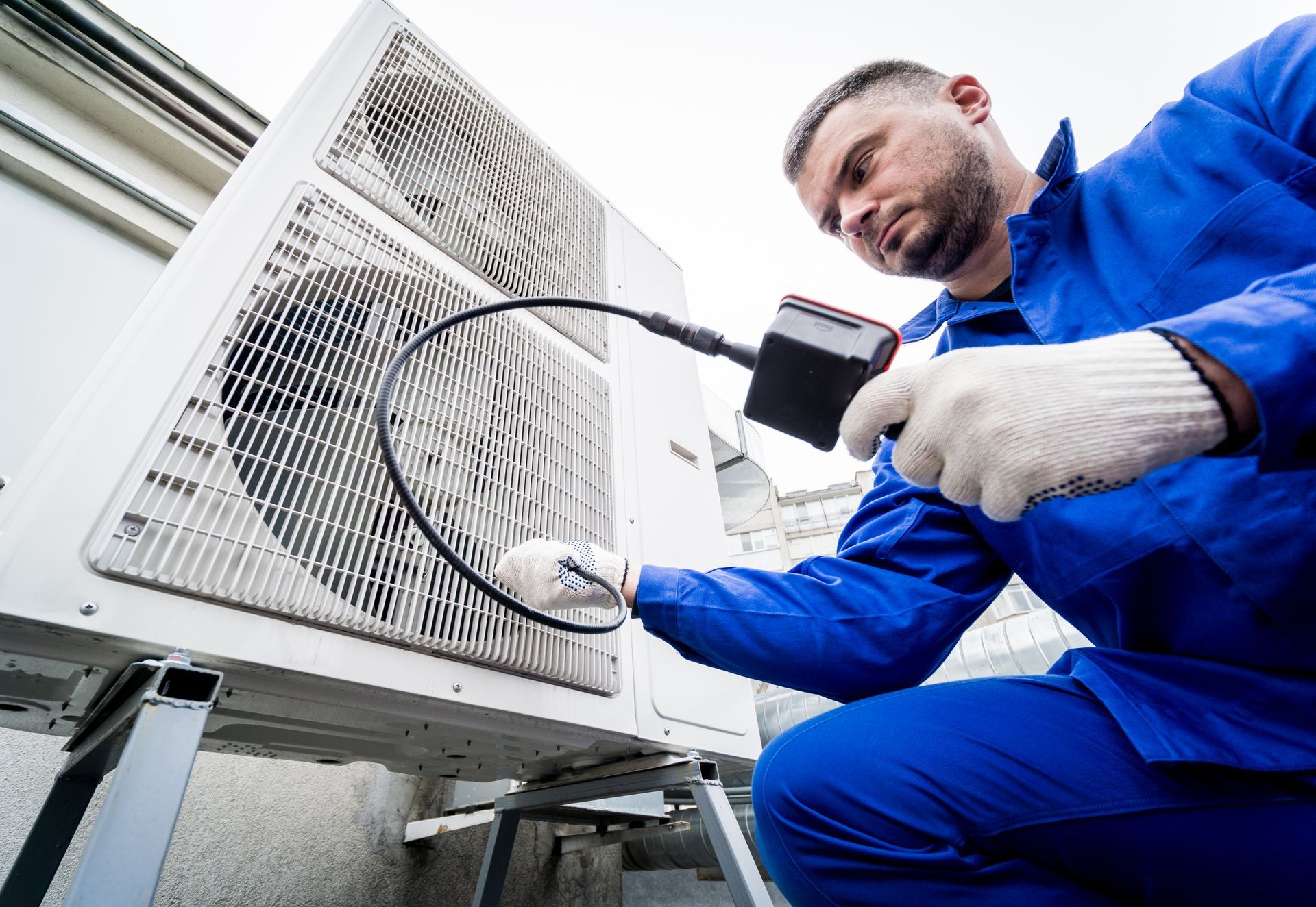 A technician in blue workwear uses a handheld diagnostic tool to inspect a dirty outdoor air conditioning unit.