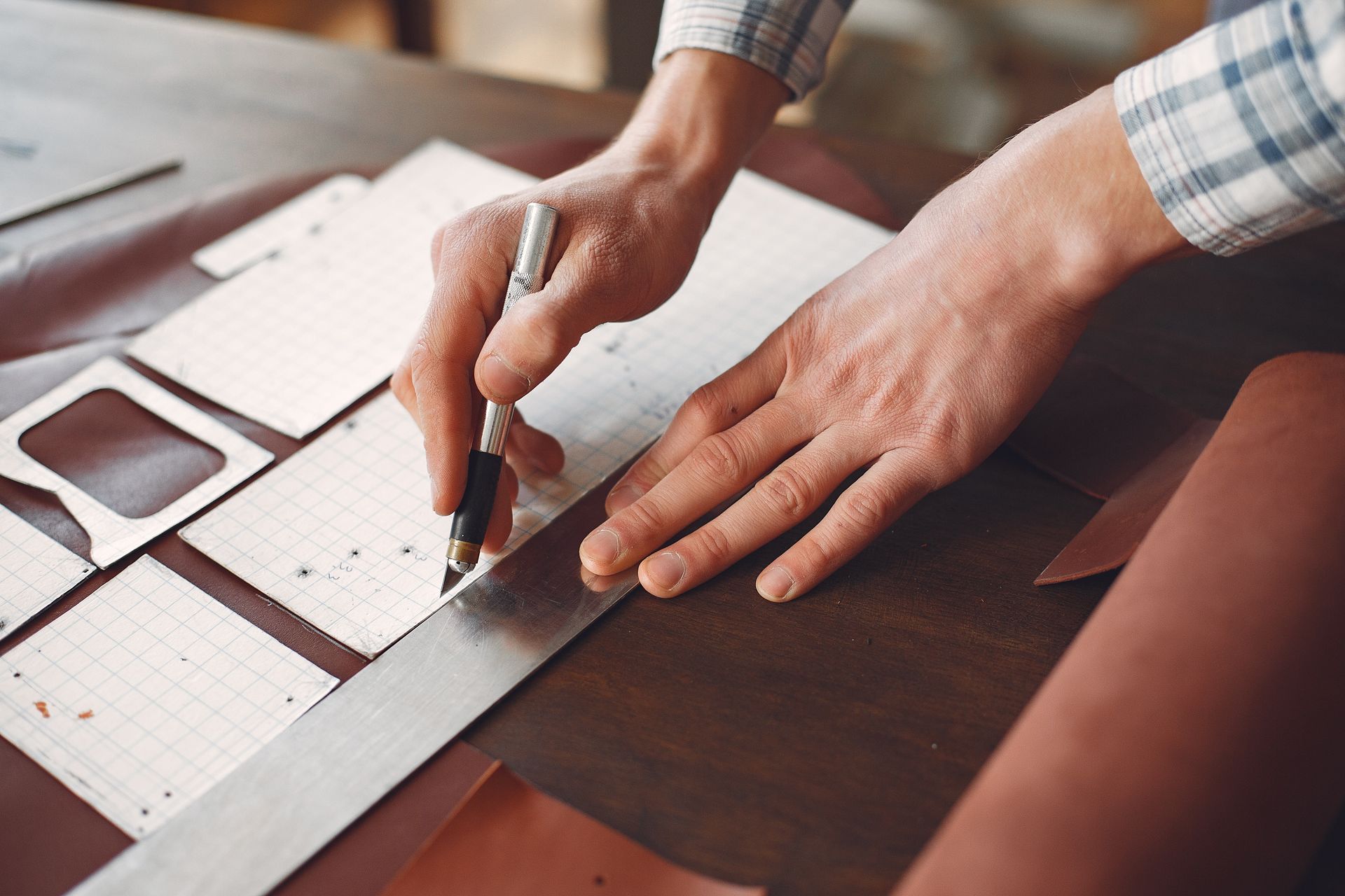 Las manos de una persona usando una regla y un bolígrafo para cortar cuero sobre una mesa.
