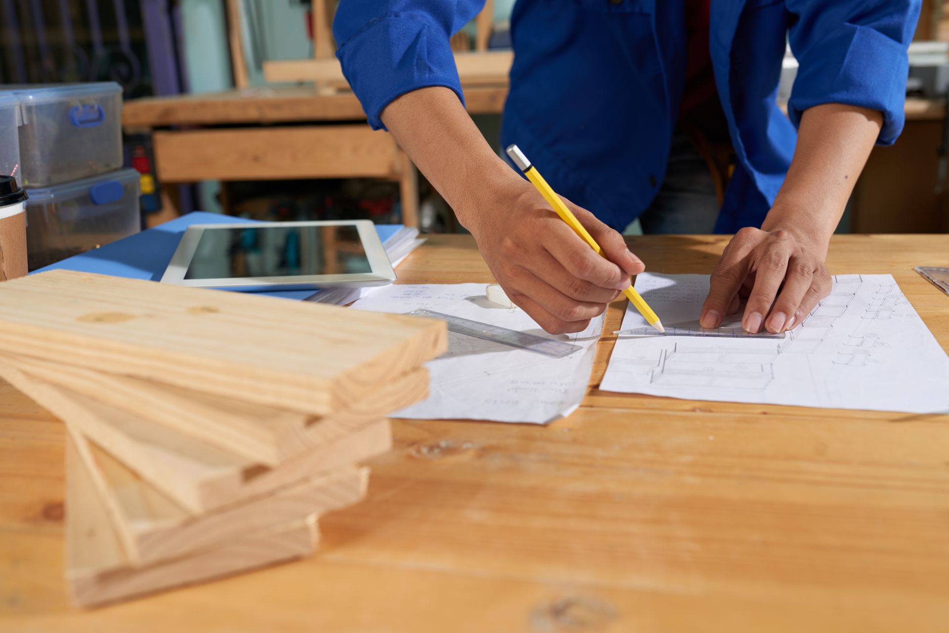Persona con chaqueta azul dibujando planos en una mesa de madera con tablones de madera; entorno de taller.