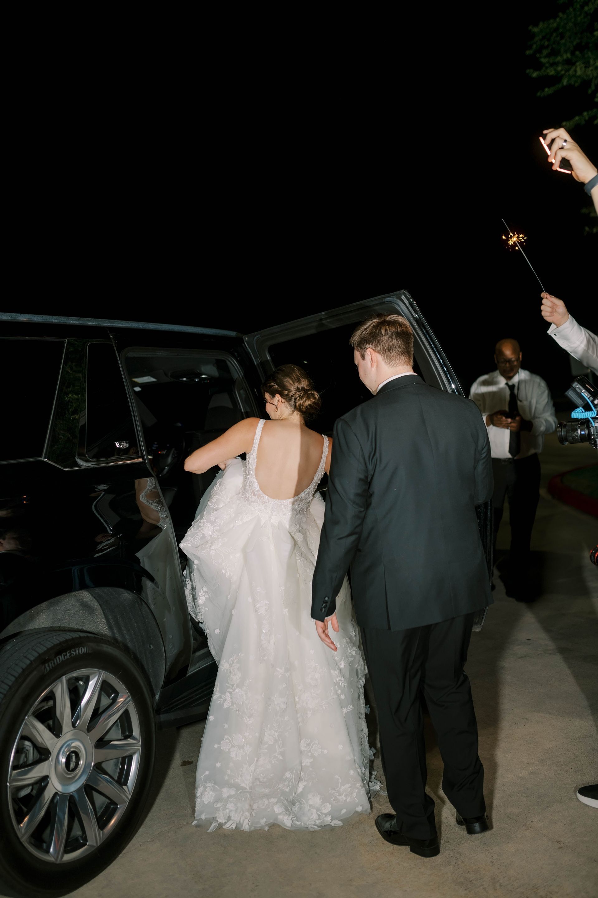 Bride and groom exiting a black car at night, dress and suit. People holding sparklers nearby.