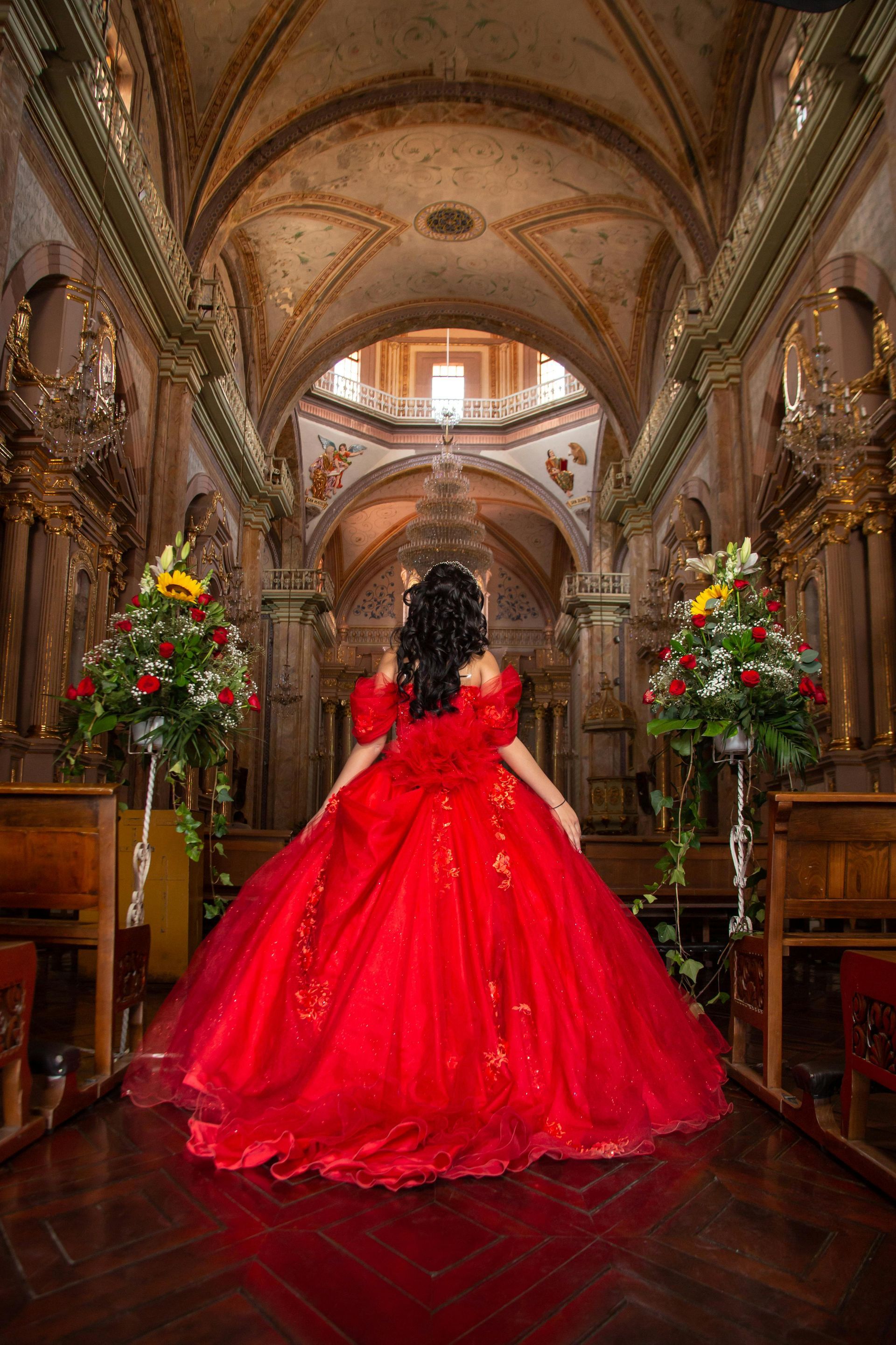 Woman in a red ballgown stands in a cathedral, back to the viewer.