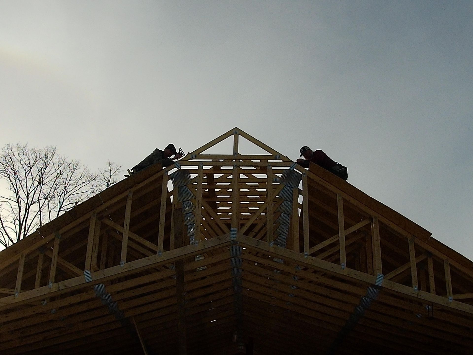 Workers on a wood truss roof frame under a cloudy sky.