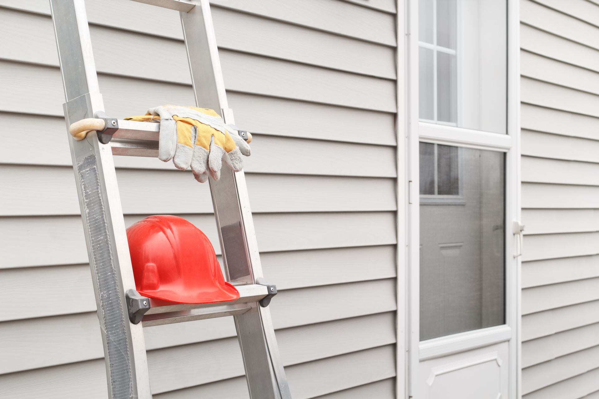 Ladder with gloves and red hard hat leaning against house siding near a door.