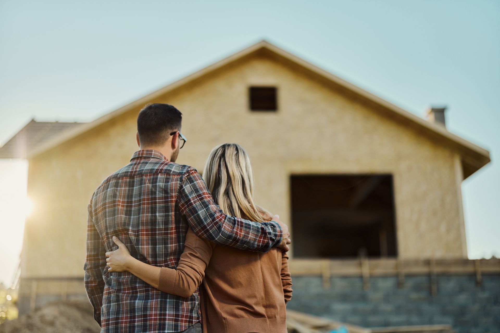Couple with arms around each other looking at a house under construction, sunlight in background.