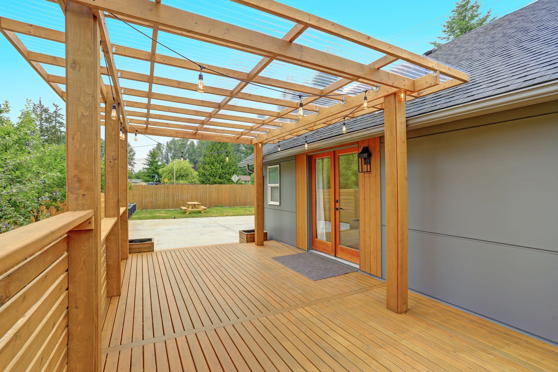 Wooden deck with pergola, leading to glass doors. A string of lights hangs overhead.