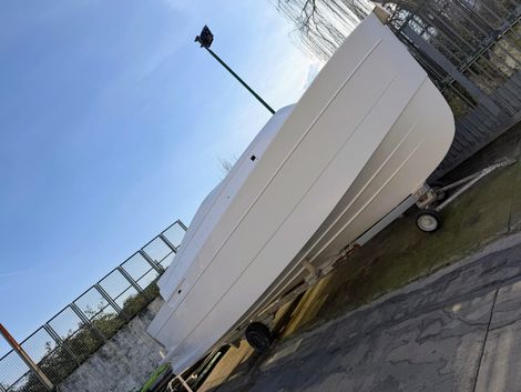 Lo scafo bianco di una barca, su un rimorchio, si trova su una piazza pavimentata con una recinzione e il cielo sullo sfondo.