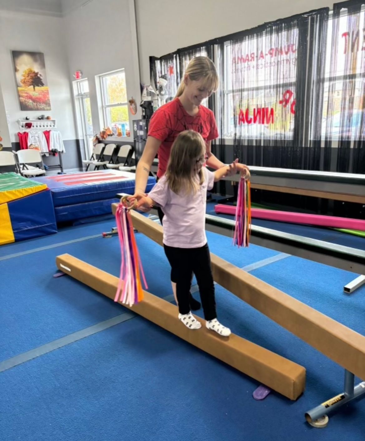 A woman is helping a little girl balance on a balance beam in a gym.